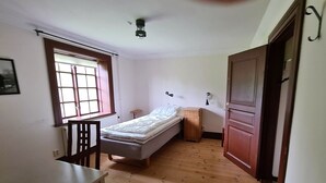 Un apartamento pequeño de una habitación con cama, una mesita, una ventana con persianas, un radiador y una puerta.