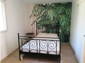 A bedroom with a bed, a small table, and a large wall mural of green leaves.