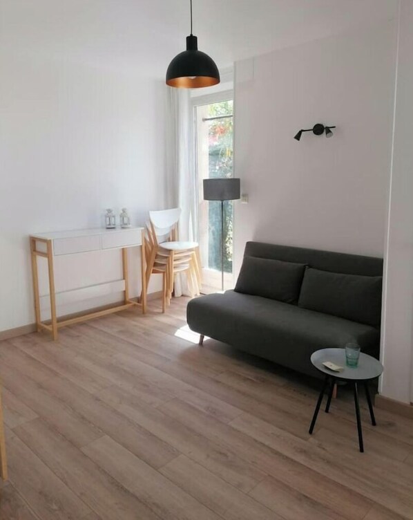 A modern living room with a grey sofa, a small round table, a dining area with a white table and chairs, and a large window with a view of greenery.