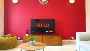 Ett vardagsrum med en röd vägg, en klocka, en Netflix-logotyp, ett träskåp, ett runt soffbord, en krukväxt, en gul sittpuff och en grå soffa.
