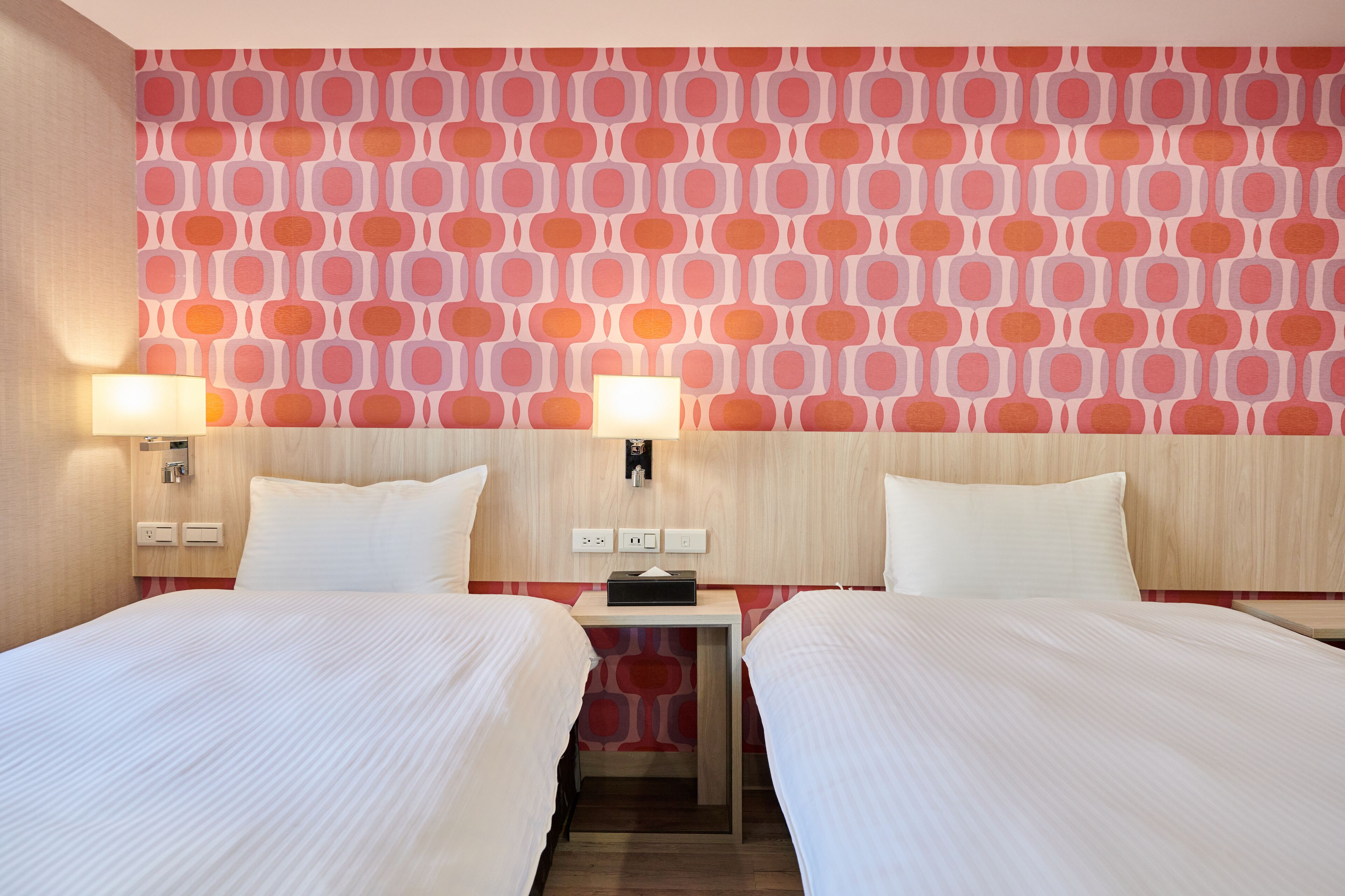 A hotel room with two beds, a red patterned wall, and a small bedside table with a lamp.