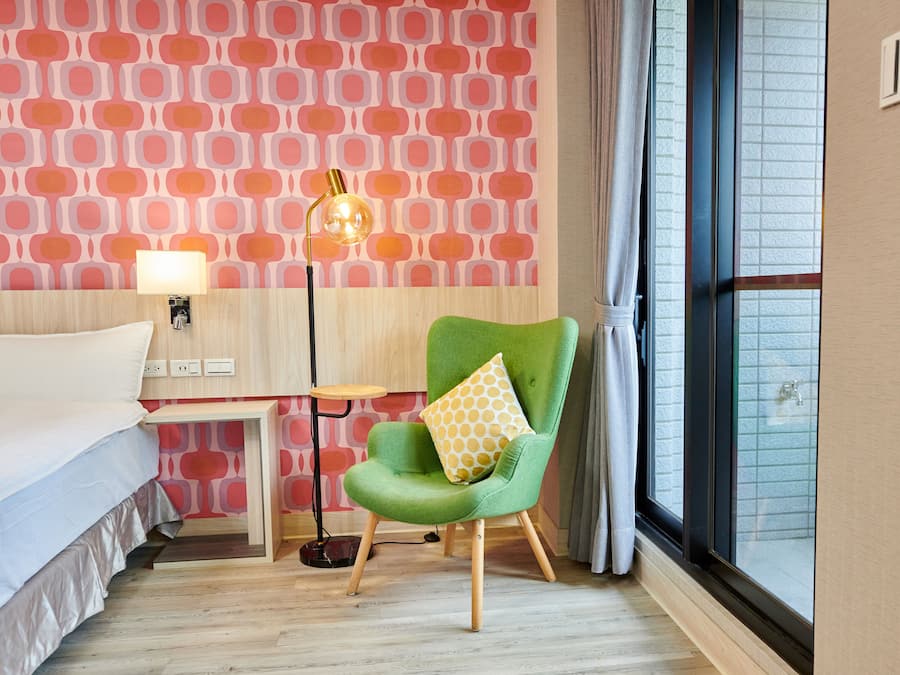 Et hotelværelse med en seng, en grøn lænestol med gul pude, en gulvlampe og et vindue med gardiner.