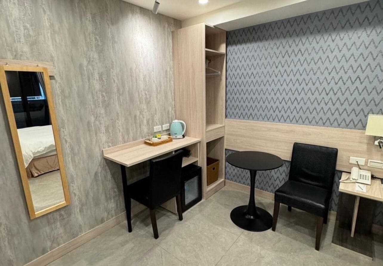 Habitación de hotel compacta con escritorio de madera, una mesita redonda pequeña, una silla negra, un armario y un espejo.