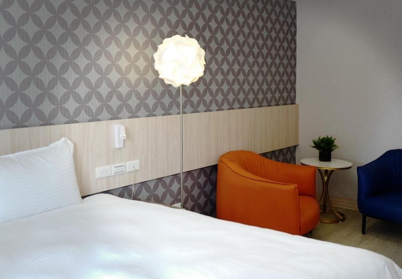 Une chambre d’hôtel avec un lit, un fauteuil orange, une chaise bleue, une petite table ronde avec une plante, et un mur orné d’un motif géométrique.
