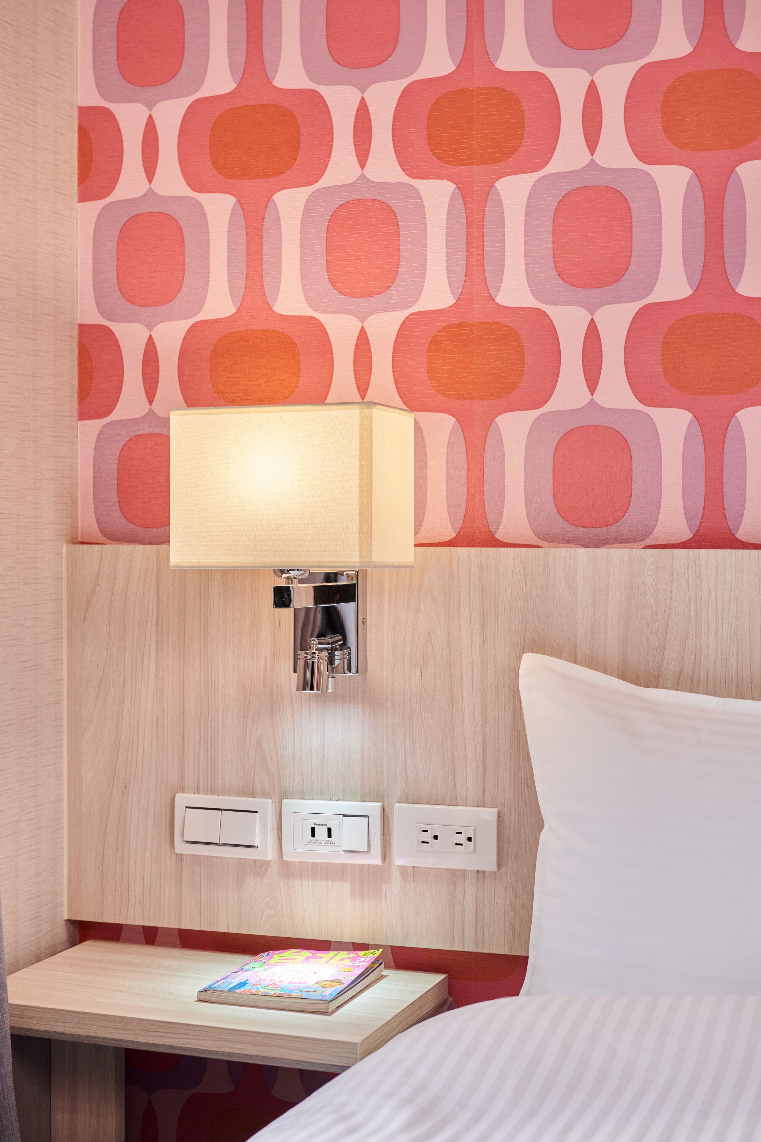 Una habitación de hotel con cabecera de madera, una mesita de noche con lámpara, un enchufe y una revista sobre la mesa.