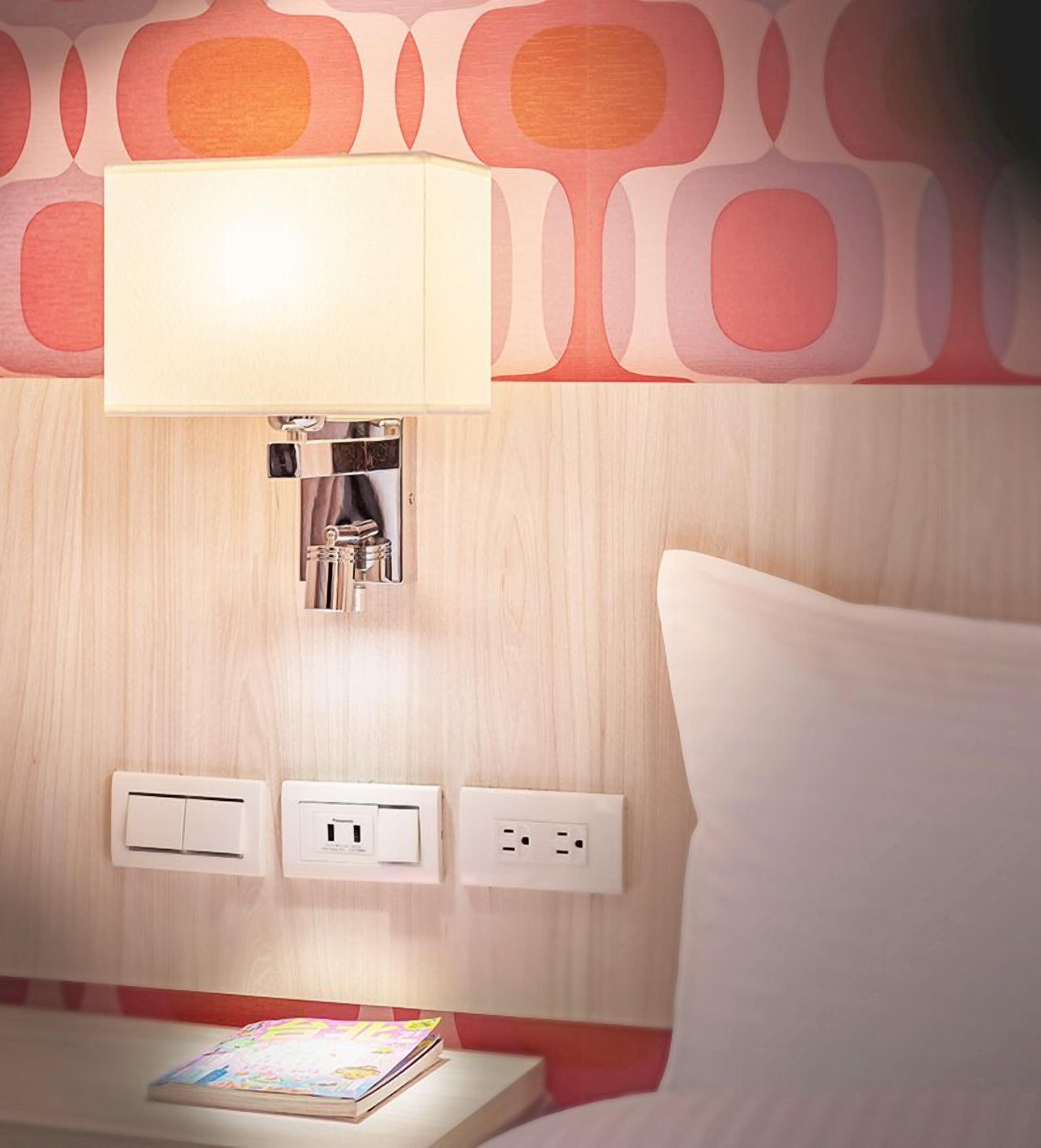 A hotel room with a bedside lamp, power outlets, and a magazine on the nightstand.