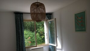 Ein Schlafzimmer mit großem Fenster, einem Bett mit gemusterten Betttextilien, einem Heizkörper und einer Hängeleuchte.