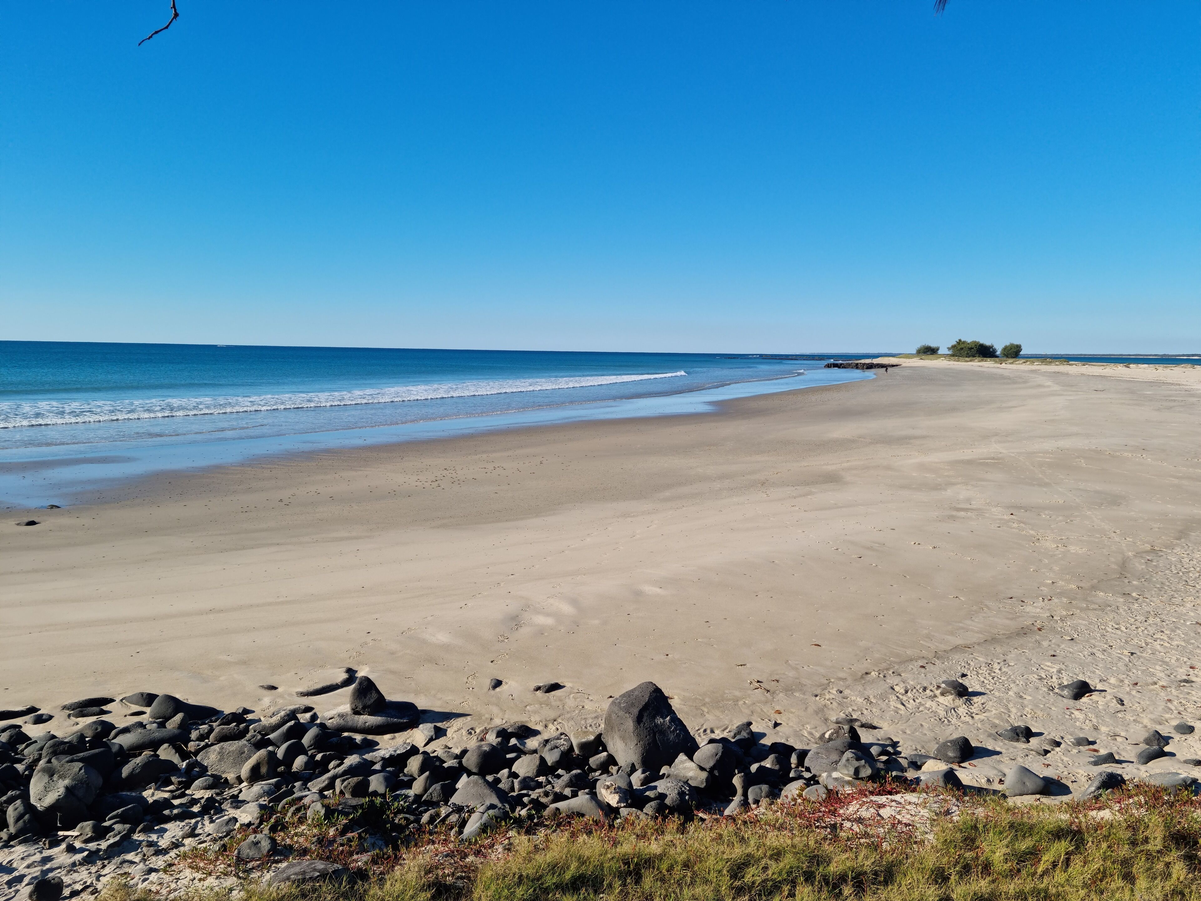 A wide sandy beach with a rocky foreground and a clear blue sky.