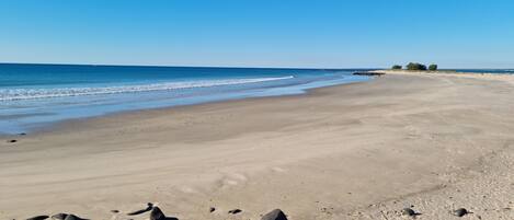 A wide sandy beach with a rocky foreground and a clear blue sky.