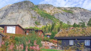 Un chalet de montagne avec un toit de chaume et des murs en bois, entouré d’une végétation luxuriante et adossé à un paysage montagneux rocheux.