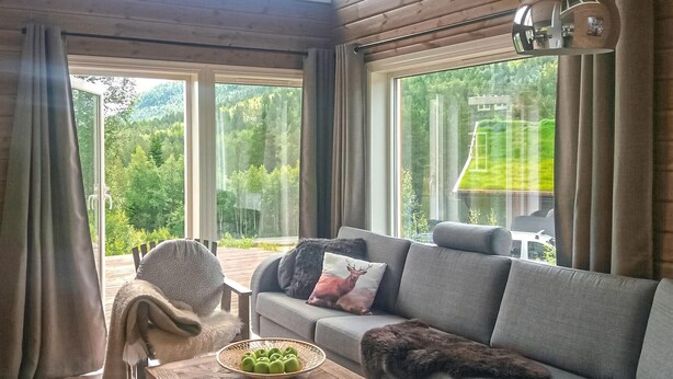 Un salon chaleureux avec un canapé gris, un panier de pommes vertes et une vue sur une forêt, visible à travers de grandes fenêtres.