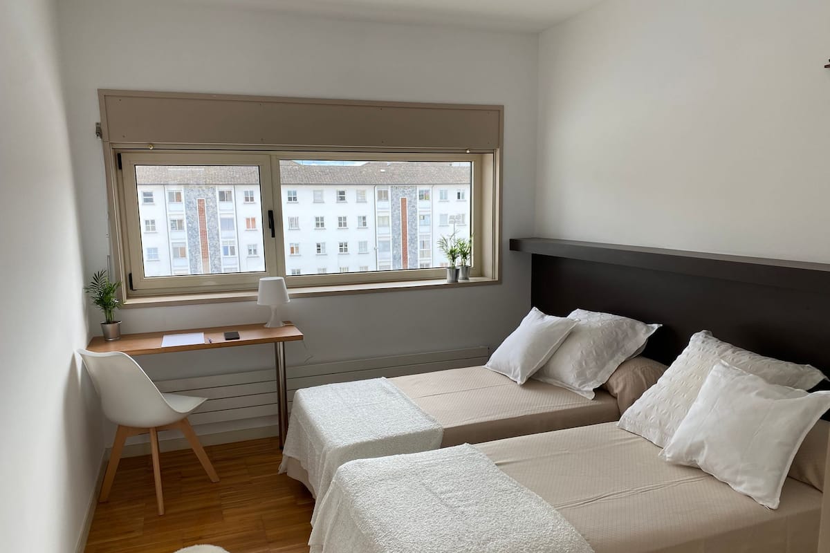A small, modern bedroom with two beds, a desk, and a chair. There is a window with a view of buildings outside.