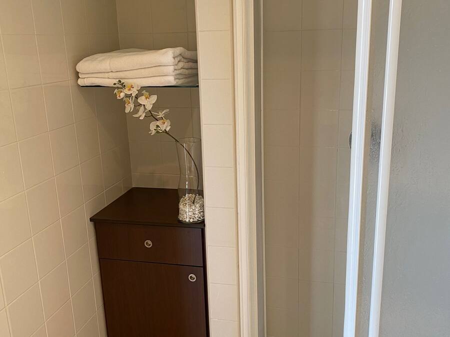 A small bathroom with a shower, a dark wooden cabinet, and a shelf with towels and an orchid vase.