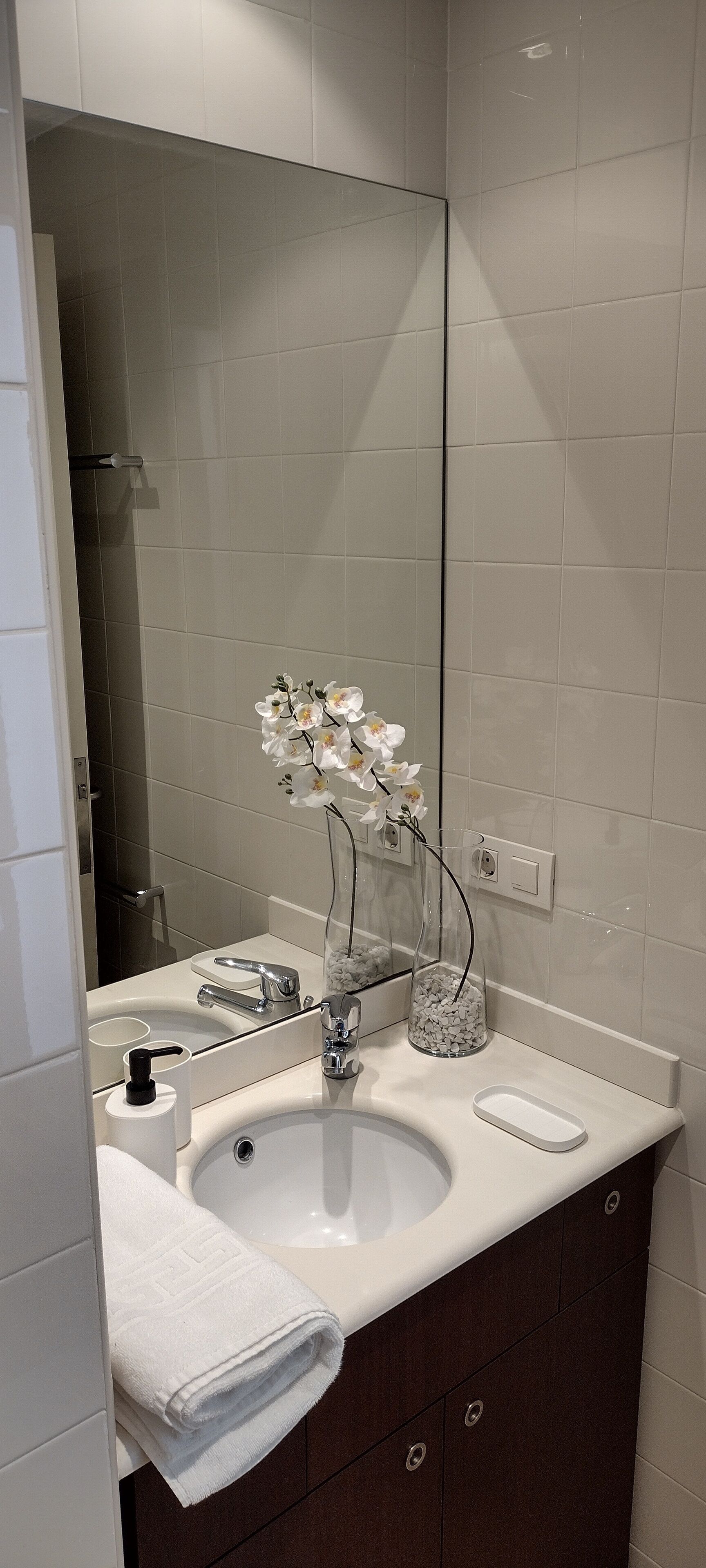 Un bagno con un lavandino bianco, due rubinetti, uno specchio e un vaso con fiori bianchi.