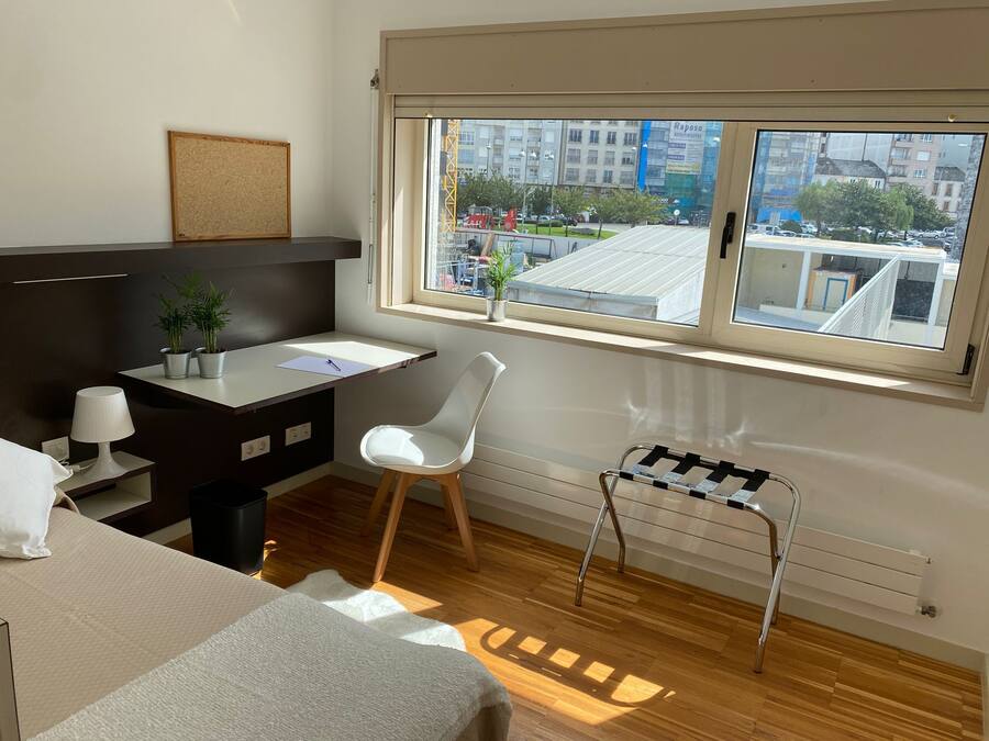 Quarto de hotel moderno com escrivaninha, cadeira e cama tipo futon. Uma janela oferece vista para a paisagem urbana.