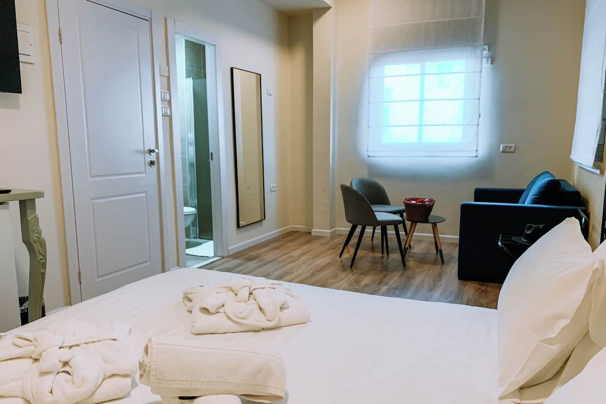 A hotel room with a bed, towels, a desk with a chair, a small table, a sofa, and a window.