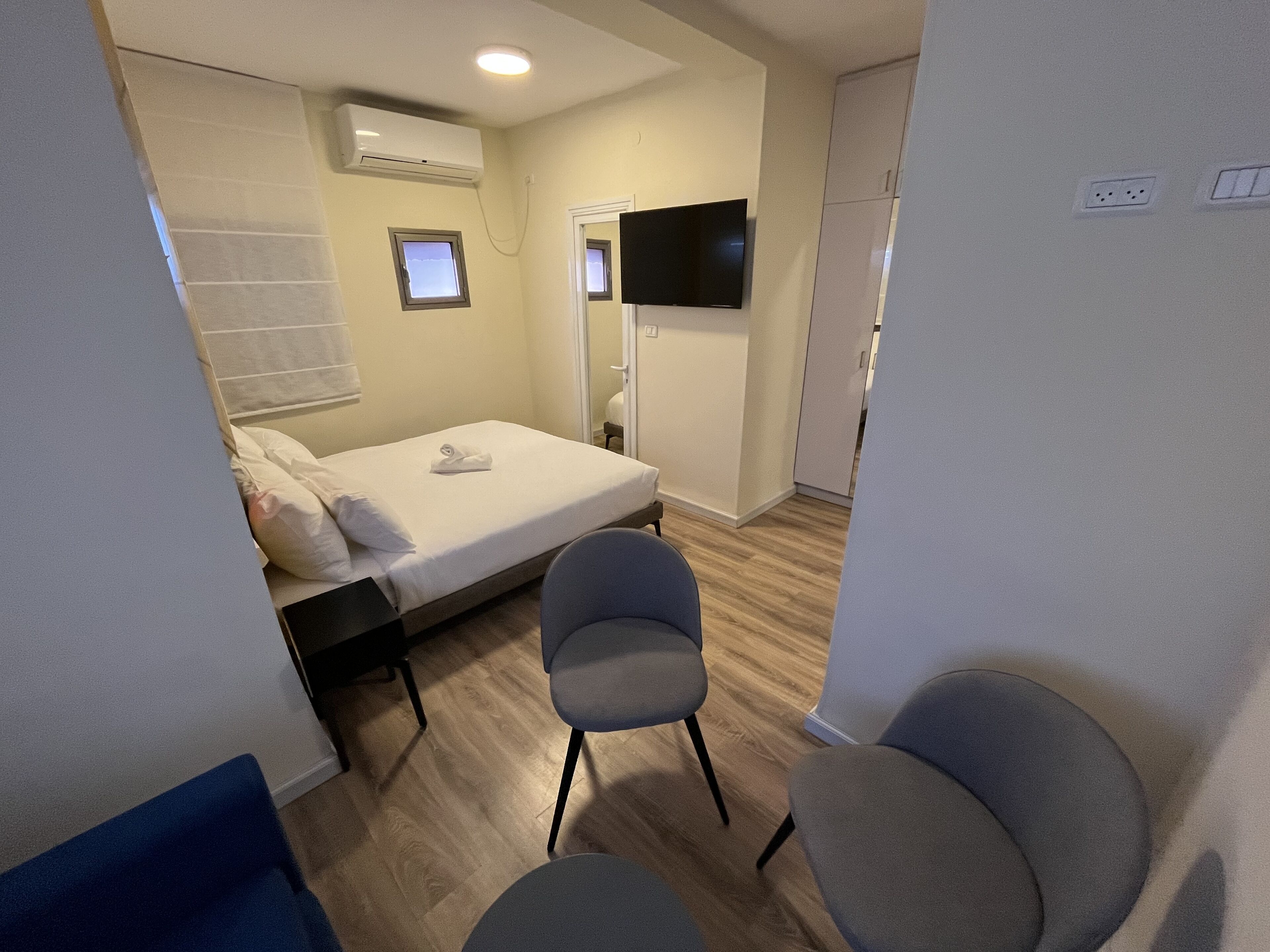 A small, well-lit room with a bed, two chairs, a TV, and an air conditioning unit.