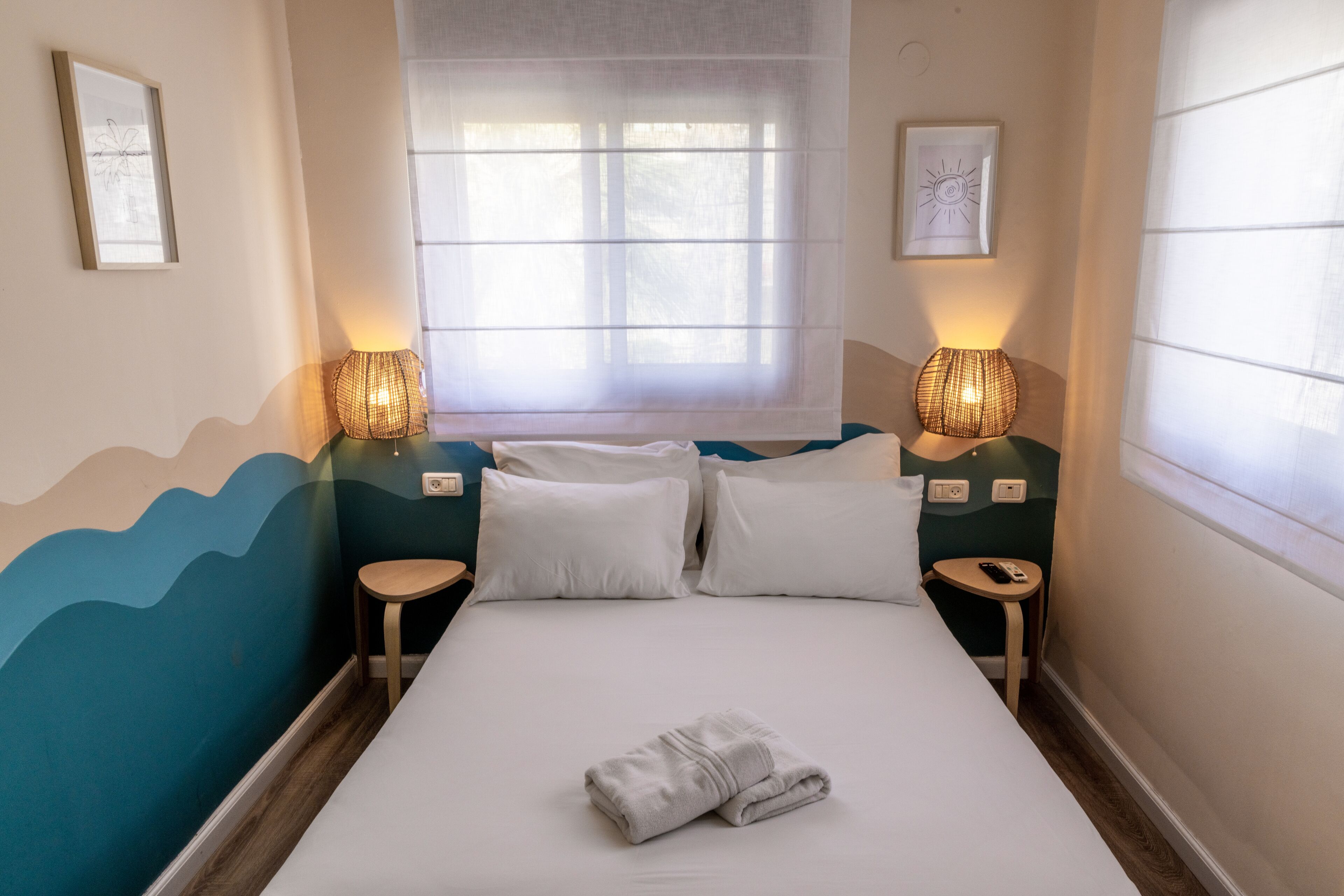 A neatly made bed with white linens, two bedside tables with lamps, a window with sheer curtains, and a wall with a wave pattern design.