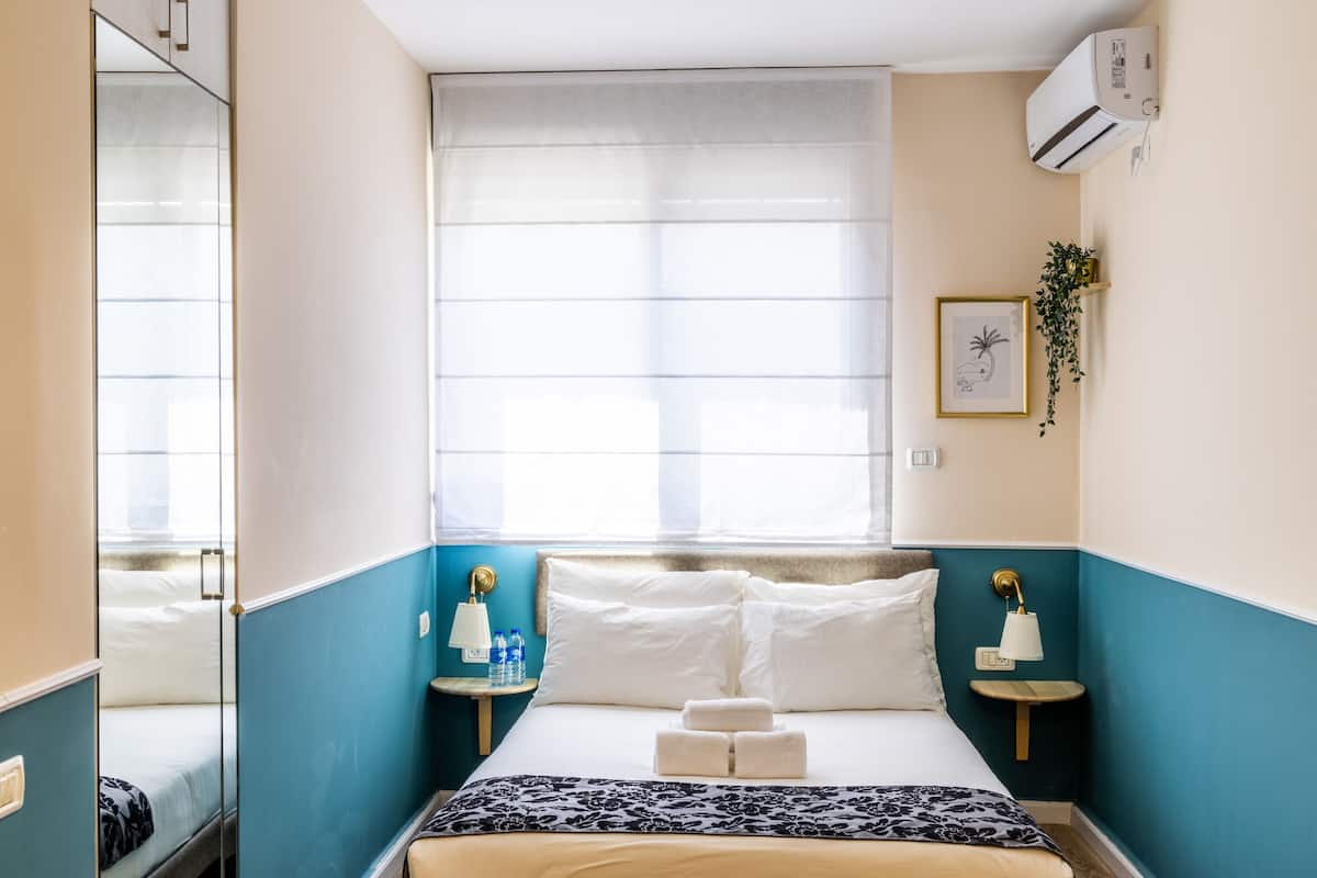 A hotel room with a bed, bedside tables, a window with curtains, an air conditioner, and a wall-mounted plant.