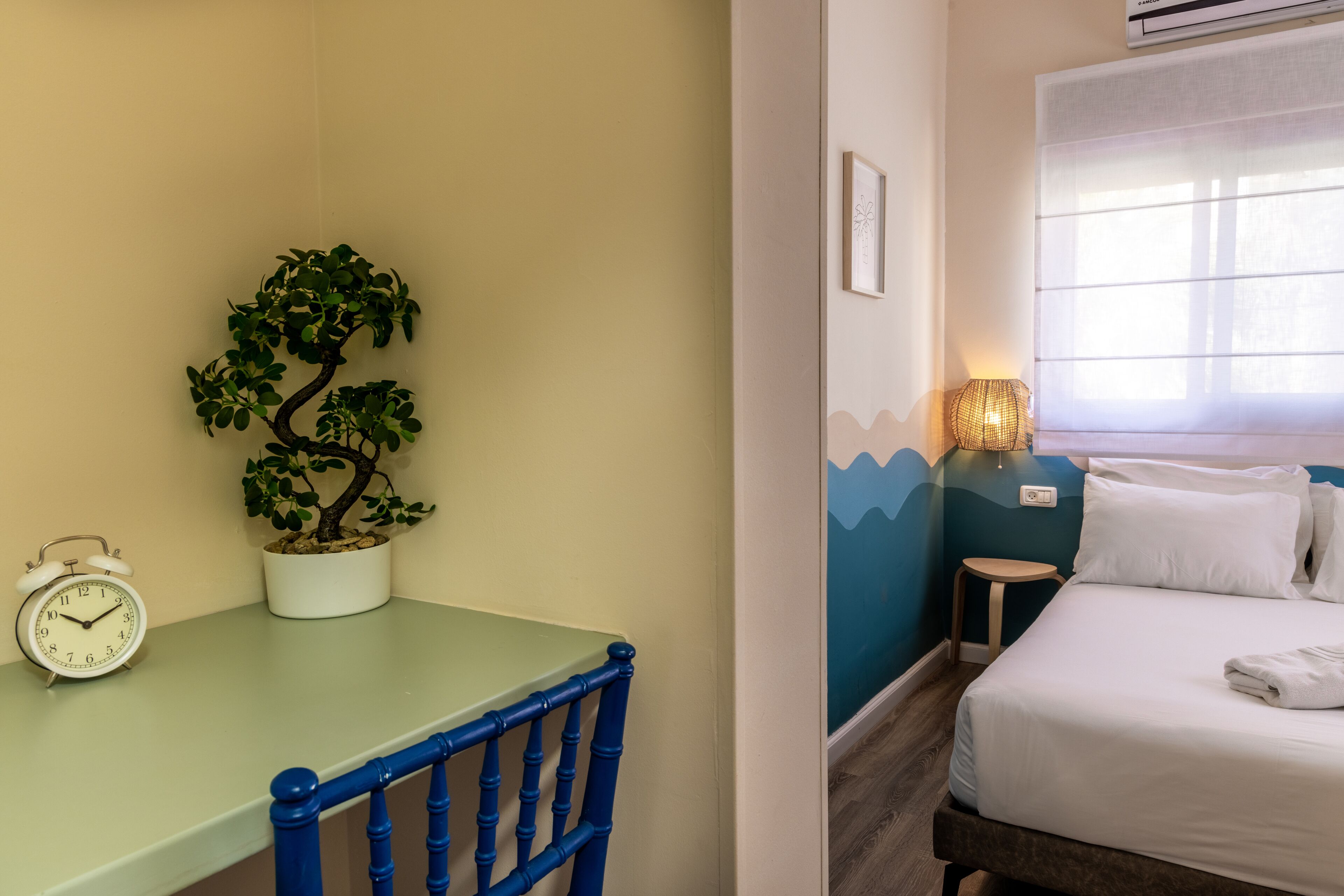 A room with a bed, a bedside table with a lamp, a potted plant, and a clock.