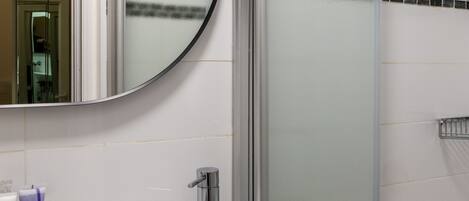 Un baño con un lavamanos blanco, un espejo redondo y una ducha con cerramiento de vidrio.
