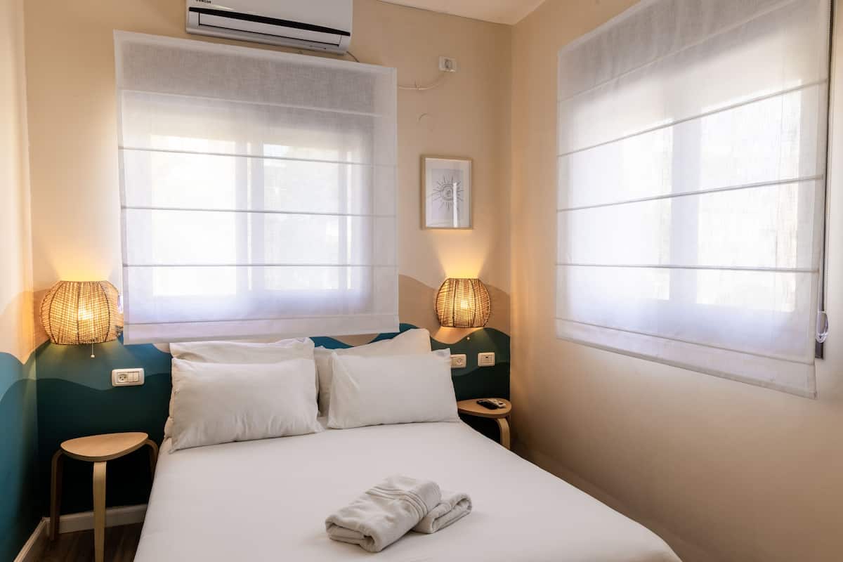 A hotel room with a bed, two bedside tables, a wall-mounted air conditioner, and two windows with sheer curtains.