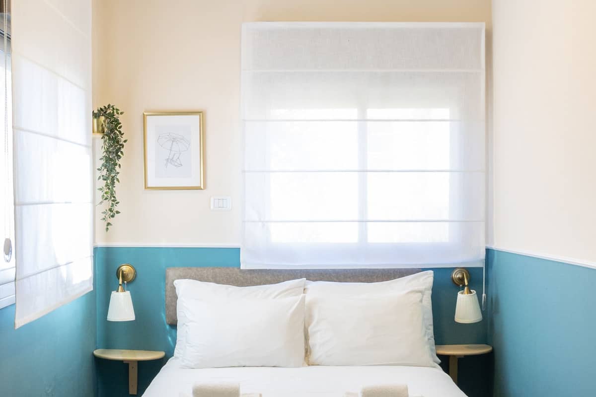 A neatly made bed with white linens and two pillows, a bedside lamp, a framed picture on the wall, and a window with a sheer curtain.