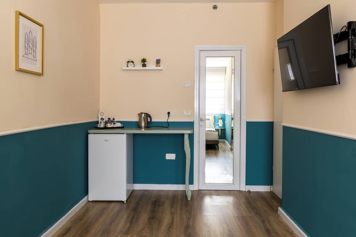 A compact kitchen with teal walls, a white refrigerator, a small countertop with a kettle, and a door leading to a bedroom.