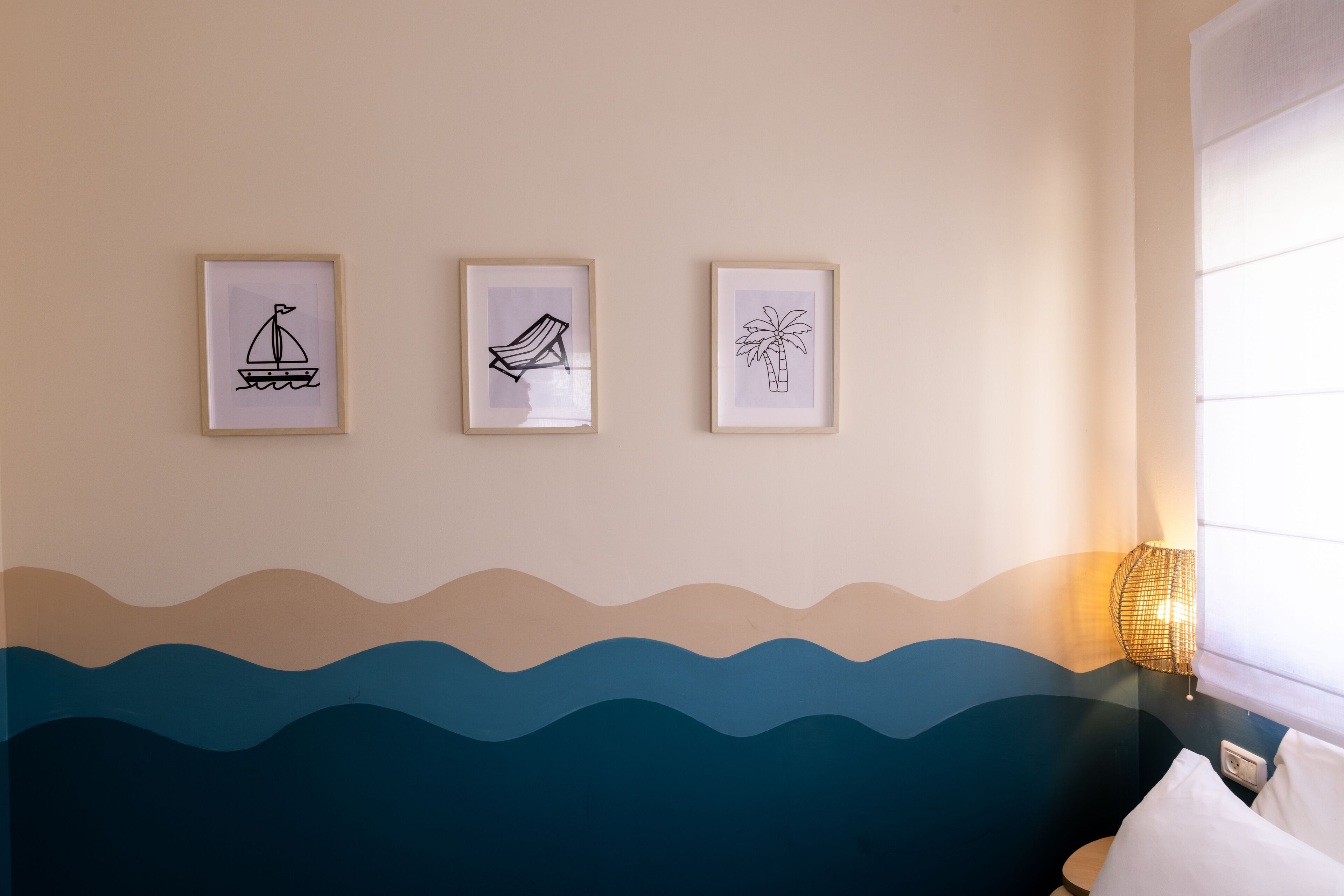 A room with a bed, a wall-mounted lamp, and three framed pictures depicting a sailboat, a beach chair, and a palm tree.