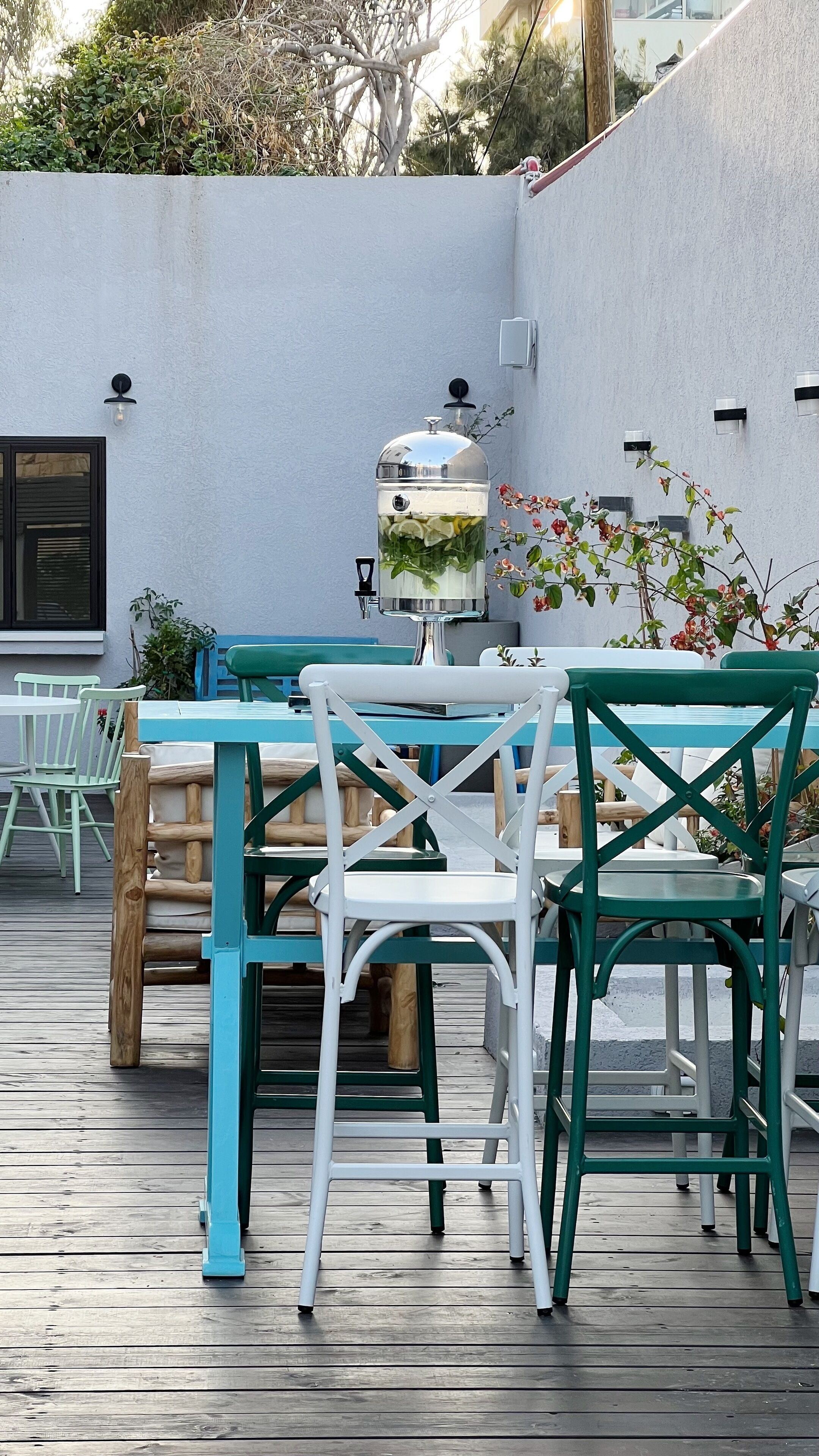An outdoor patio with a wooden table and chairs, a water feature, and a white wall with small windows.
