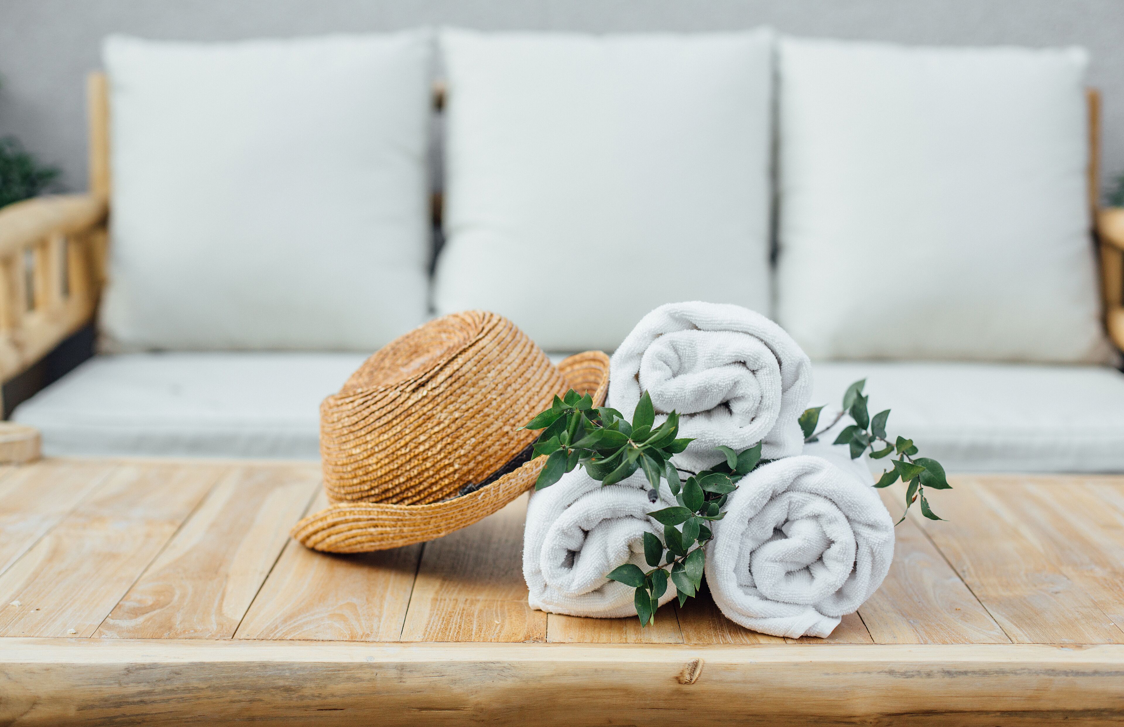 A wooden table with rolled white towels and a straw hat, set against a white sofa with cushions.