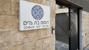 A stone wall with a sign featuring a logo and text, a metal gate, and flags in the background. - Domus Bat Galim Hotel (Haifa)