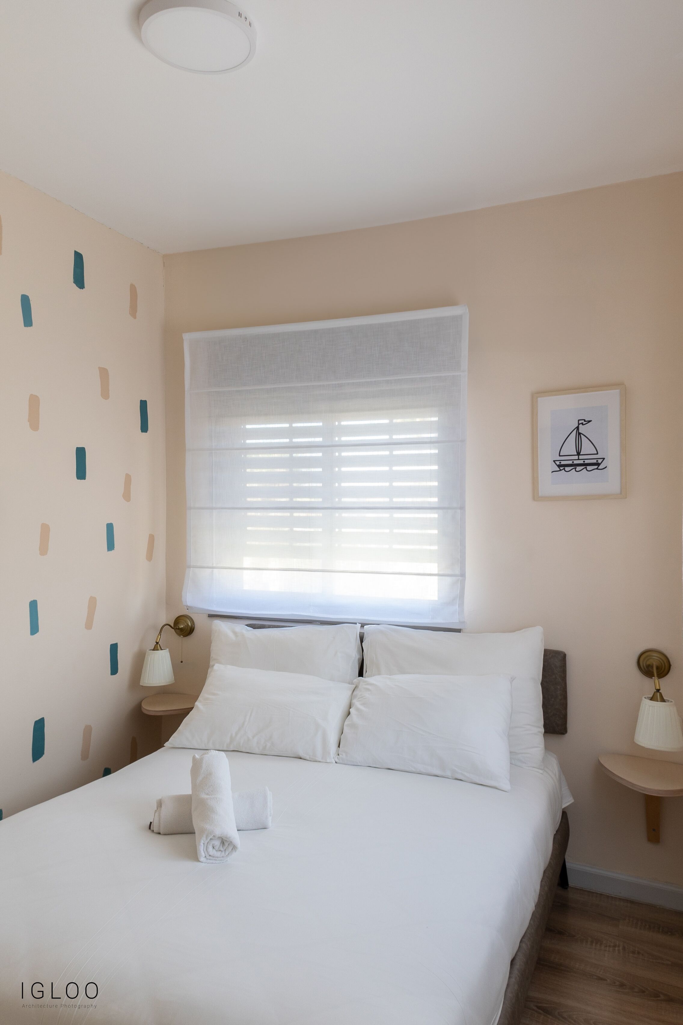 A neatly made bed with white linens and a rolled towel, a window with blinds, and a wall with abstract patterns.