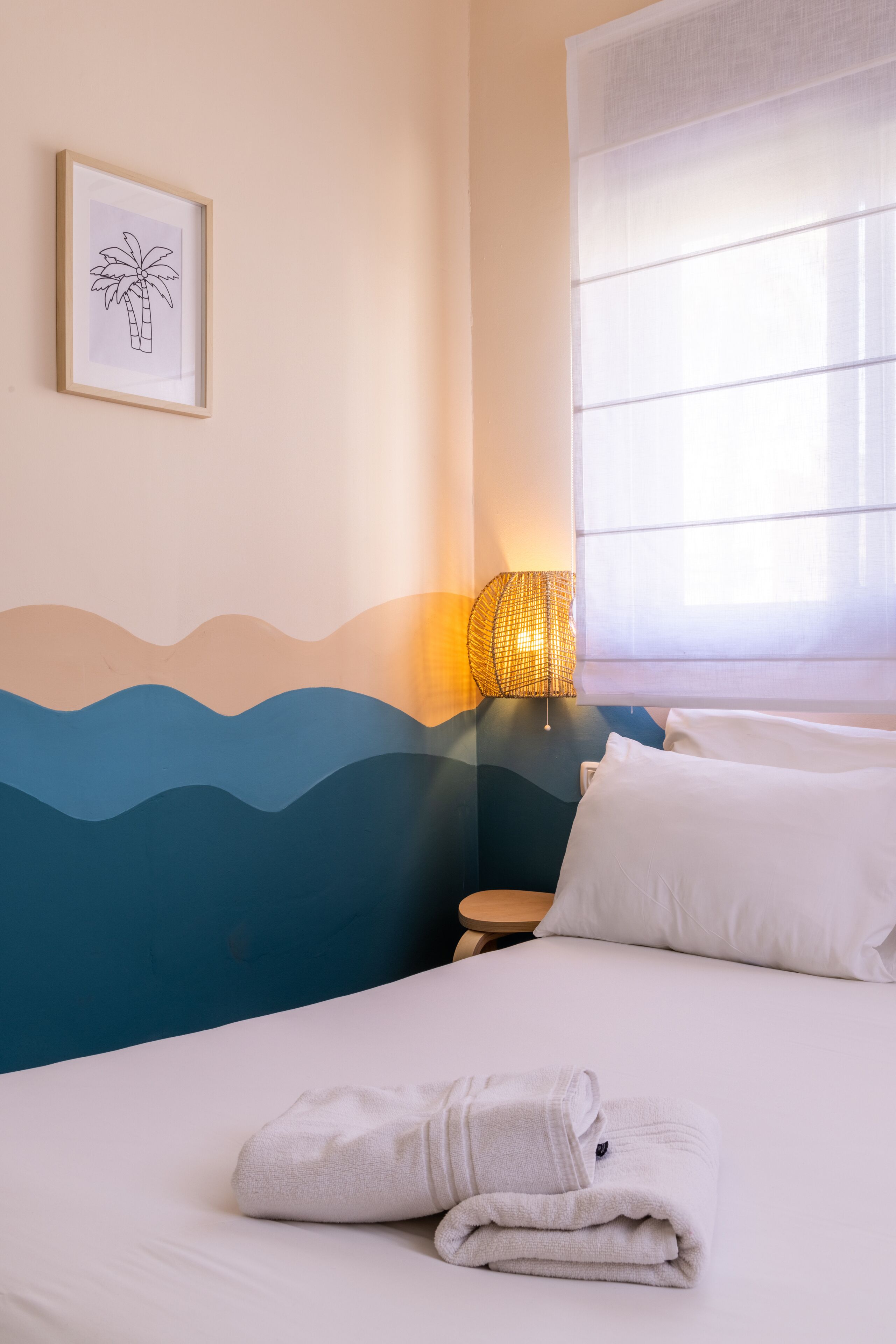 Una cama bien hecha con sábanas blancas y una toalla doblada, un cuadro en la pared de una palmera, una lámpara de noche y una ventana con cortinas traslúcidas.