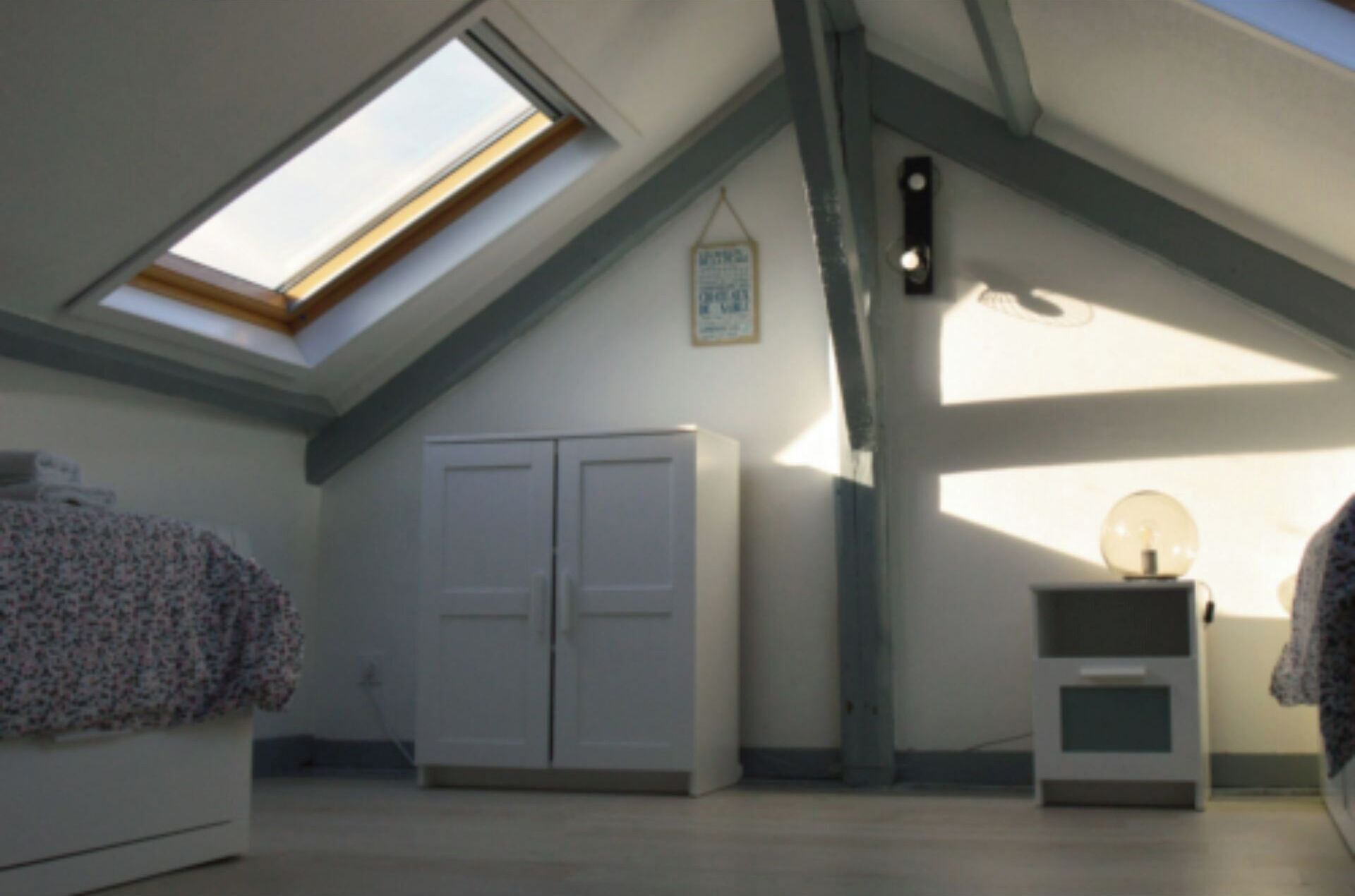 A room with a sloped ceiling, a skylight, a wardrobe, a small table with a lamp, and a wall-mounted light fixture.