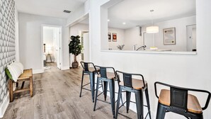 Eine moderne Küche mit großem Fenster, Spüle und Arbeitsplatte. Im Essbereich stehen drei Hocker und eine Bank.
