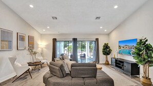 Ein modernes Wohnzimmer mit einer grauen Eckcouch, einem Flachbildfernseher und einem großen Fenster mit Blick auf einen Pool und Sitzmöglichkeiten im Freien.