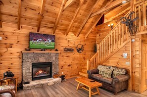 A cozy cabin interior with a stone fireplace, wooden walls, a television mounted above it, a staircase, and a leather sofa. - 3 BR/2.5 BA NEAR PARKWAY, GAME ROOM, FIREPLACE, HOT TUB,REAL LOG CABIN,PETS OK! (Pigeon Forge)