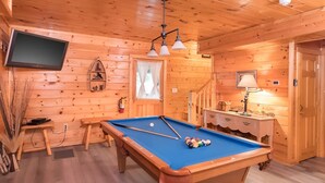 Interior de uma cabana de madeira com mesa de bilhar, televisão fixada na parede e escada que leva a outro andar.