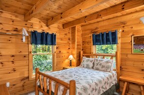 A wooden cabin room with a bed, a nightstand, a lamp, and a window with curtains. - 3 BR/2.5 BA NEAR PARKWAY, GAME ROOM, FIREPLACE, HOT TUB,REAL LOG CABIN,PETS OK! (Pigeon Forge)