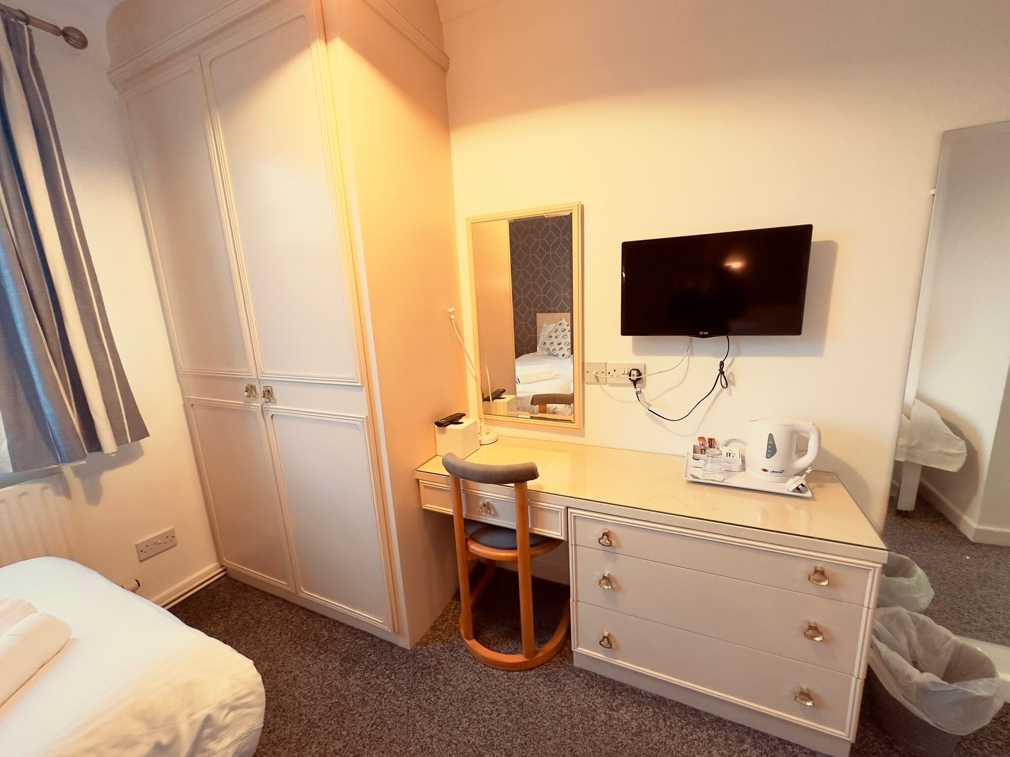 Habitación de hotel con cama, escritorio con silla, televisión, espejo y hervidor de agua.