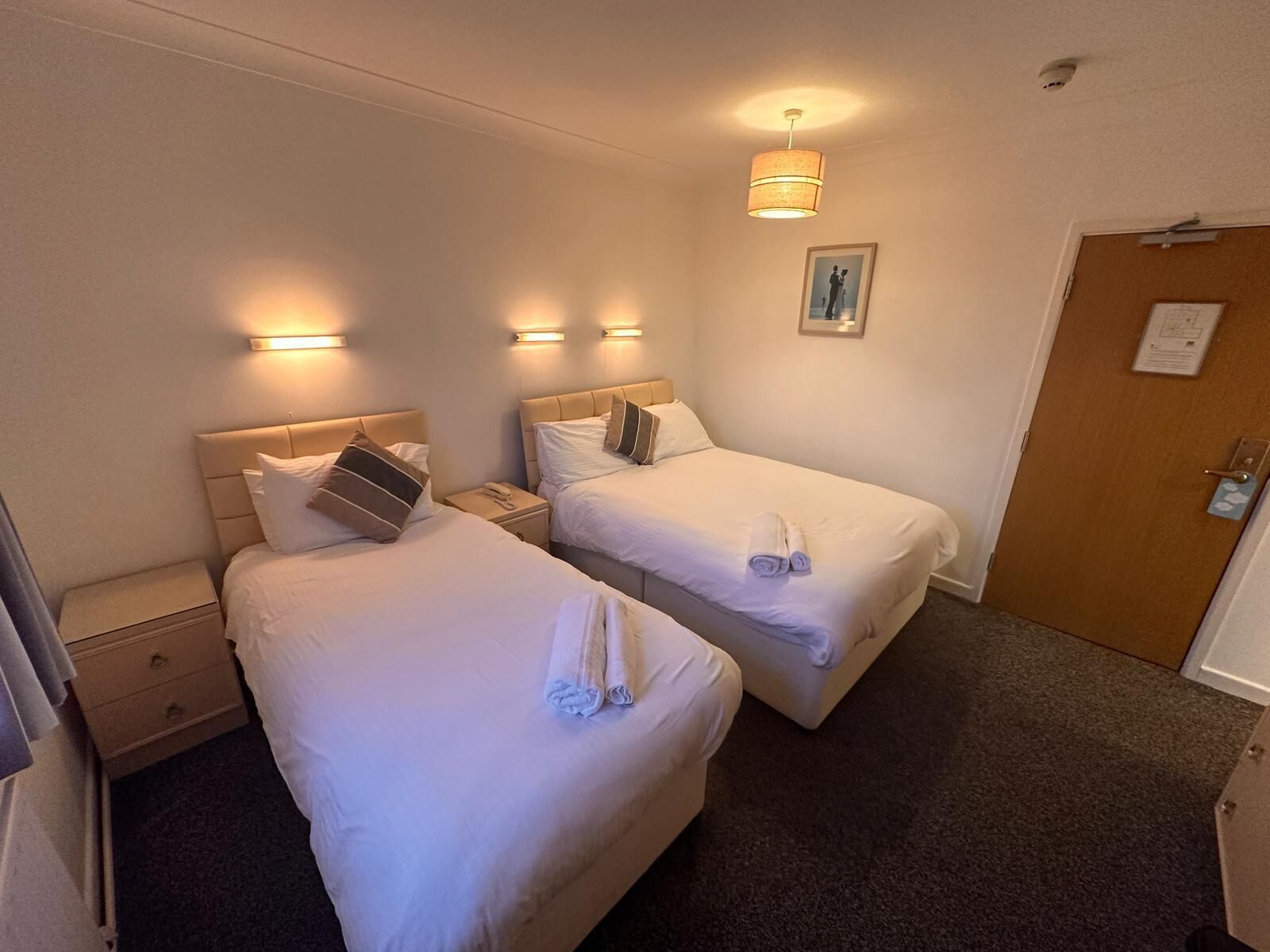 Habitación de hotel con dos camas, cada una con cabecero, mesitas de noche y lámparas. Se aprecia una puerta con cerradura y un ventanal pequeño.