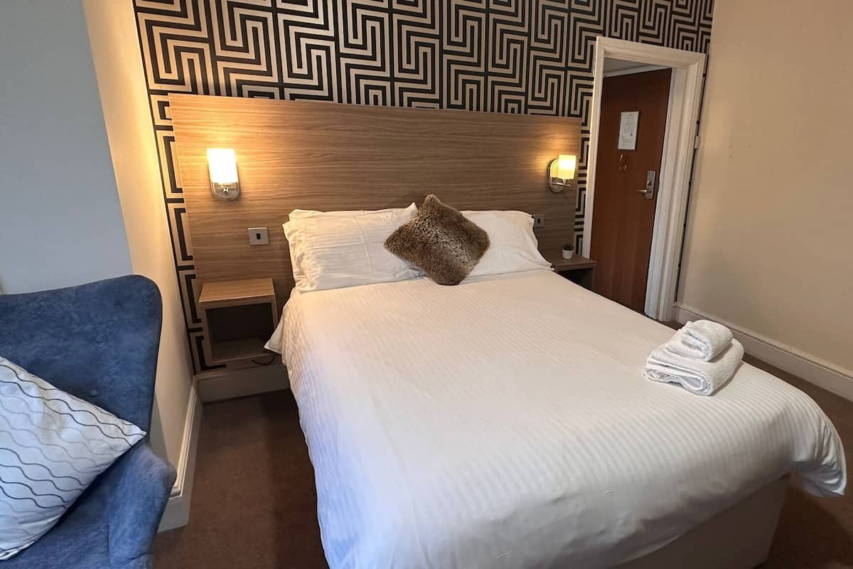 Un lit bien fait, avec du linge de lit blanc, un coussin marron et une serviette pliée posée sur le côté. On y trouve une table de chevet en bois avec une lampe, une petite plante en pot et un luminaire fixé au mur. Une chaise bleue est placée à gauche du lit.