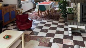 Zona pranzo con pavimento a scacchi, tavolo con tovaglia rossa e una sedia rossa. Si vede una finestra con tende, un armadio e un camino.