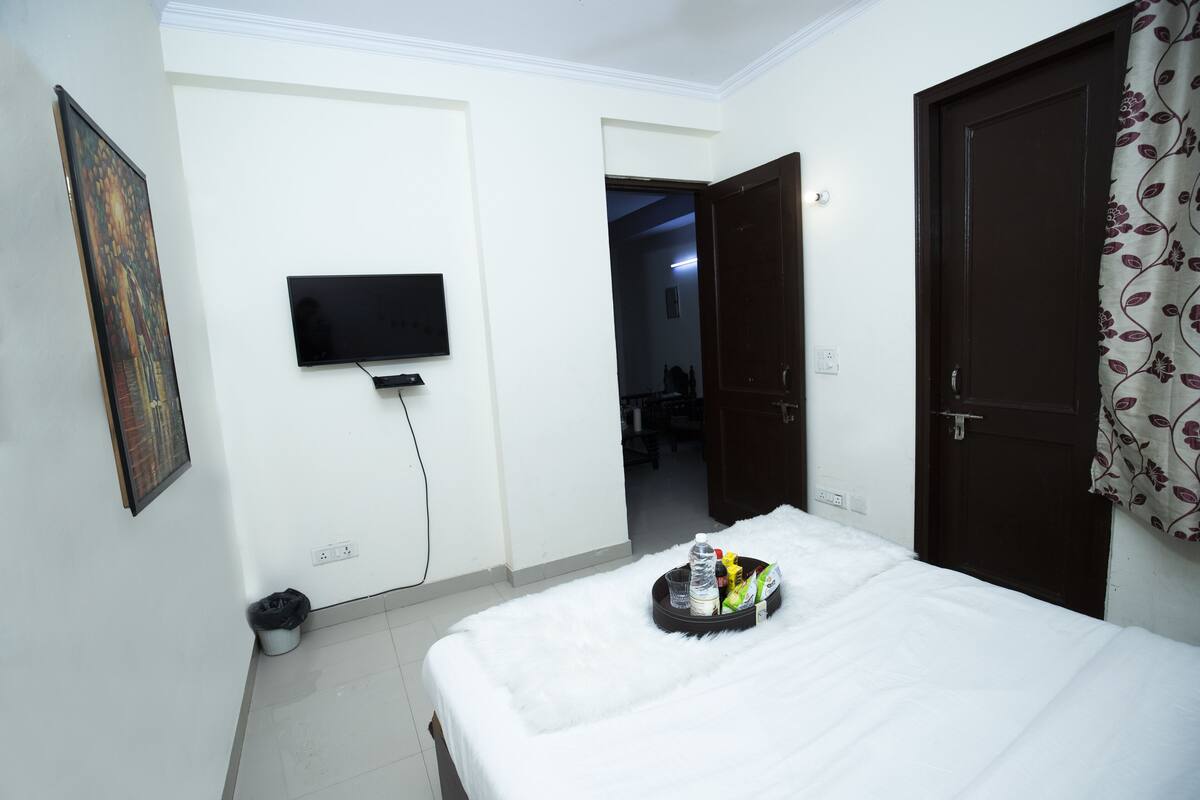 A hotel room with a bed, a television mounted on the wall, a door, and a hallway leading to another room.