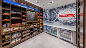 A snack and sundries section with shelves stocked with various packaged snacks, a refrigerated display case, and a wall mural advertising treats. - Hampton Inn Columbus Downtown, GA (Columbus)