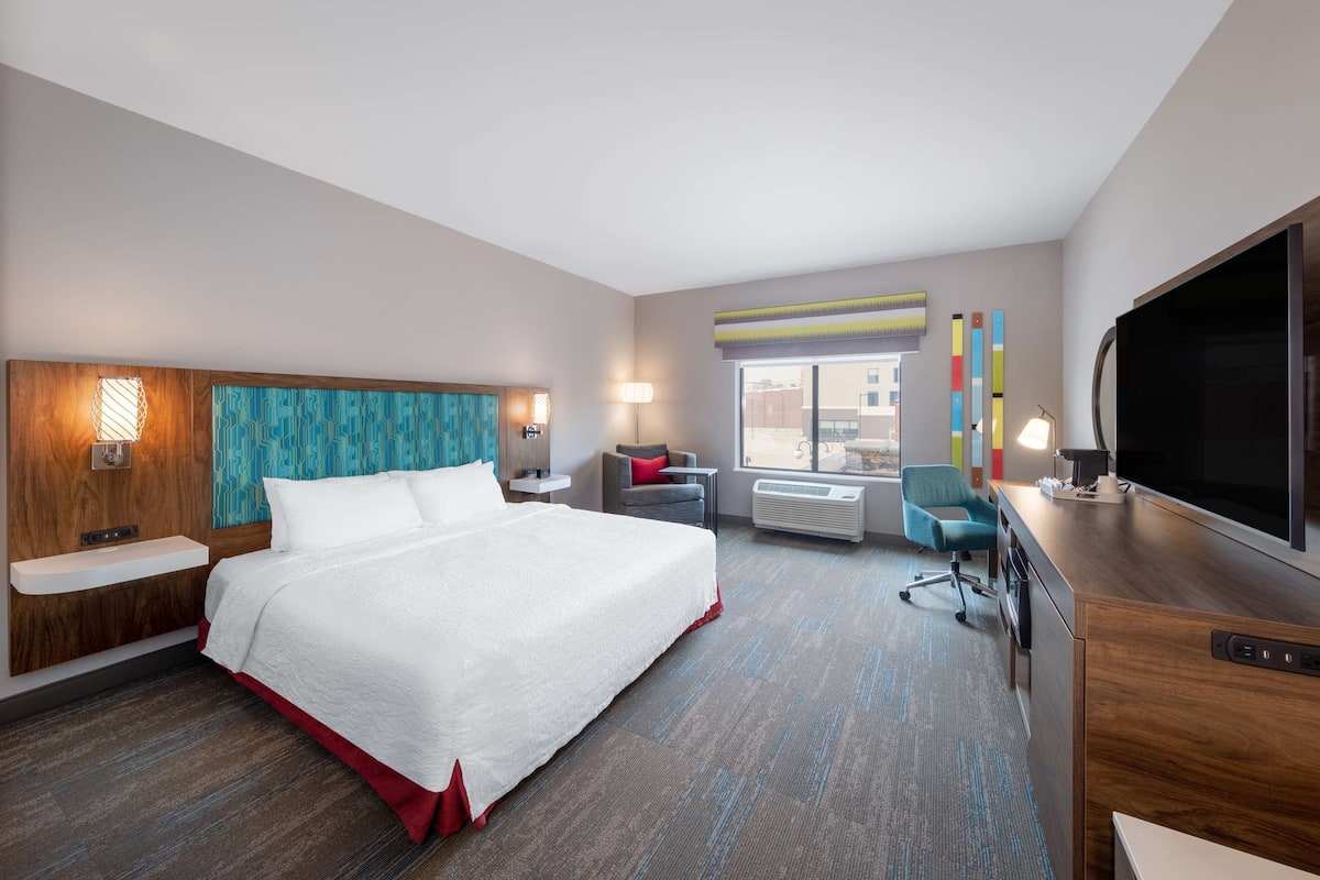A hotel room with a large bed, a desk with a flat-screen TV, a chair, a desk lamp, a window with a view of buildings, and a wall with a geometric pattern.