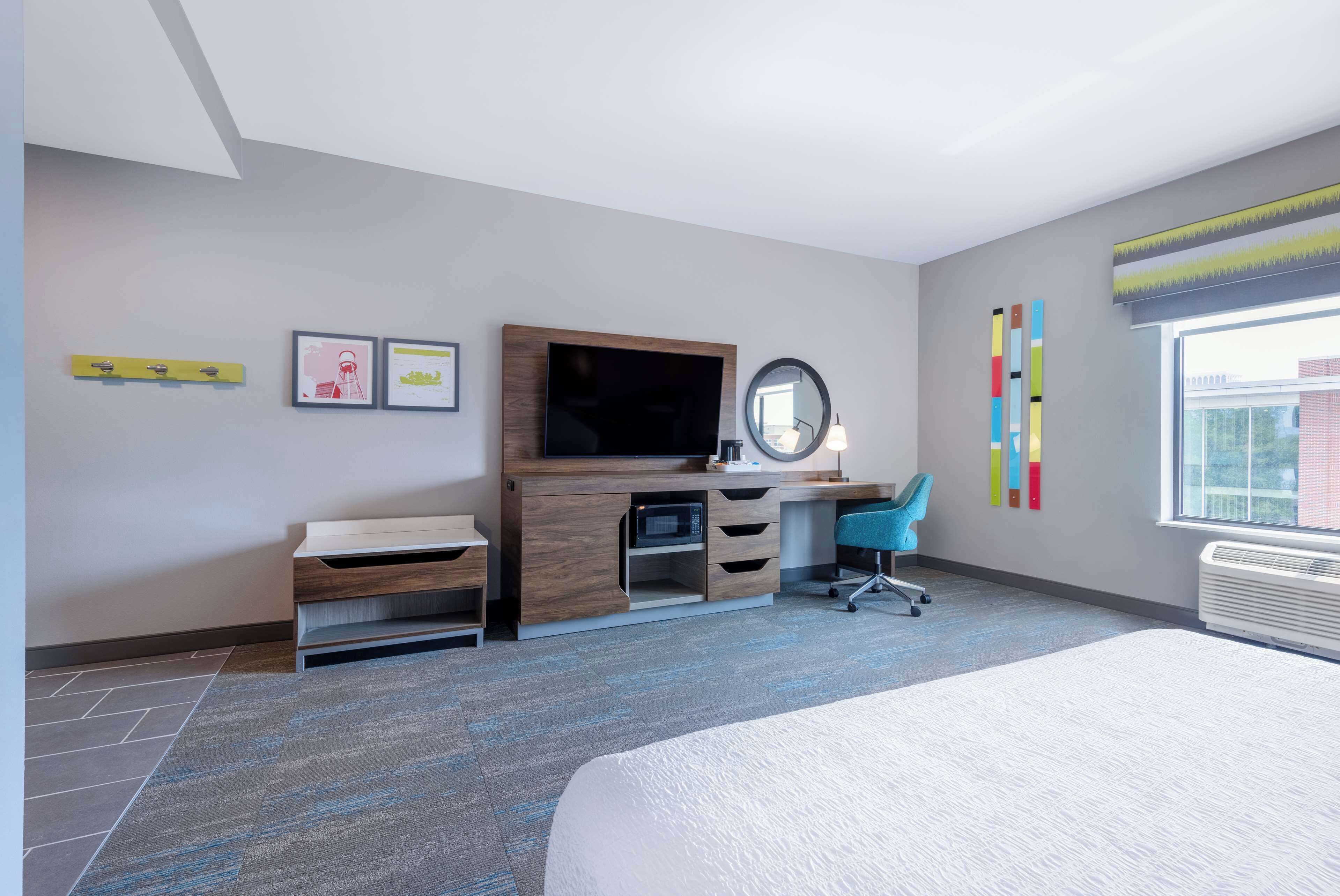 A modern hotel room with a bed, a desk with a computer, a chair, a television, and a large window.