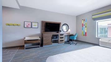 A modern hotel room with a bed, a desk with a computer, a chair, a television, and a large window.