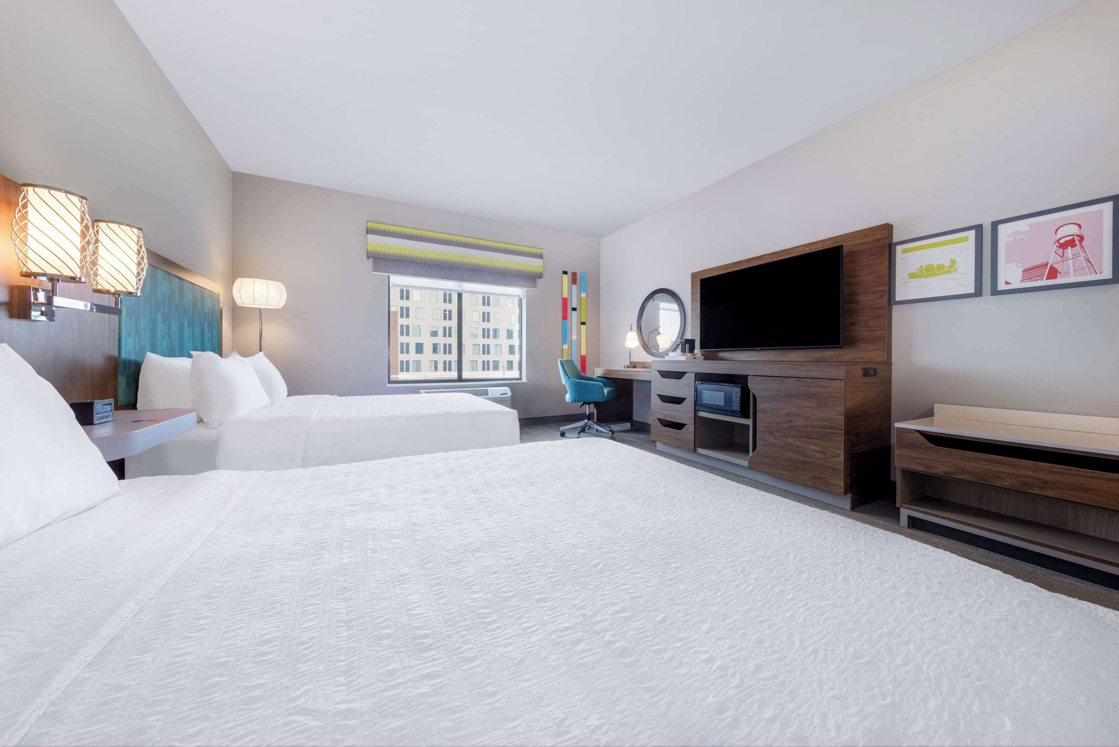 A hotel room with two beds, a desk, a chair, a TV, and a window with a view of buildings.